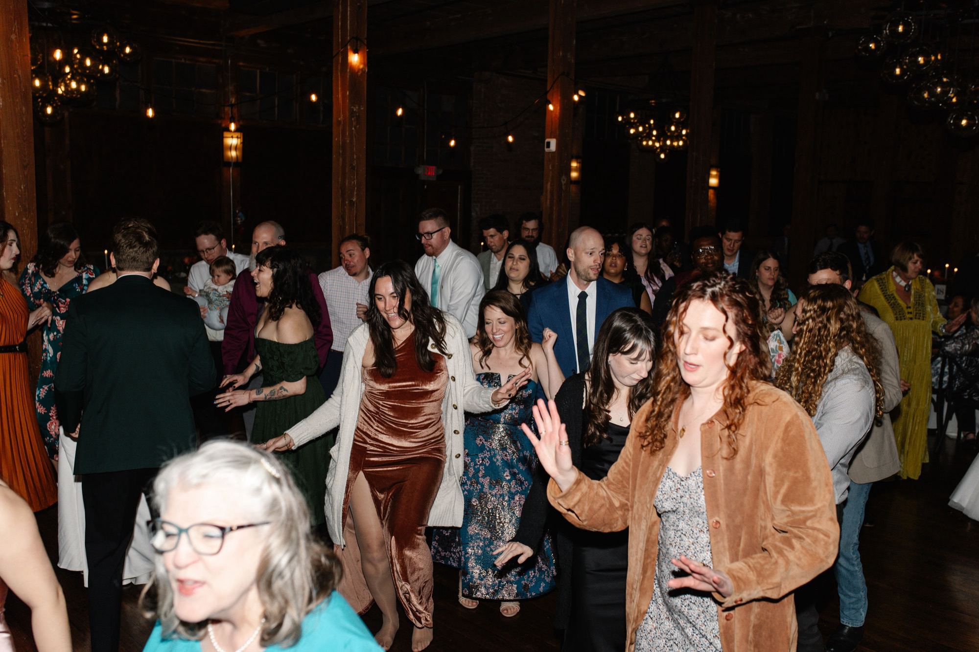 A crowd of guests dancing together on a dance floor in a wood-beamed venue under string lights