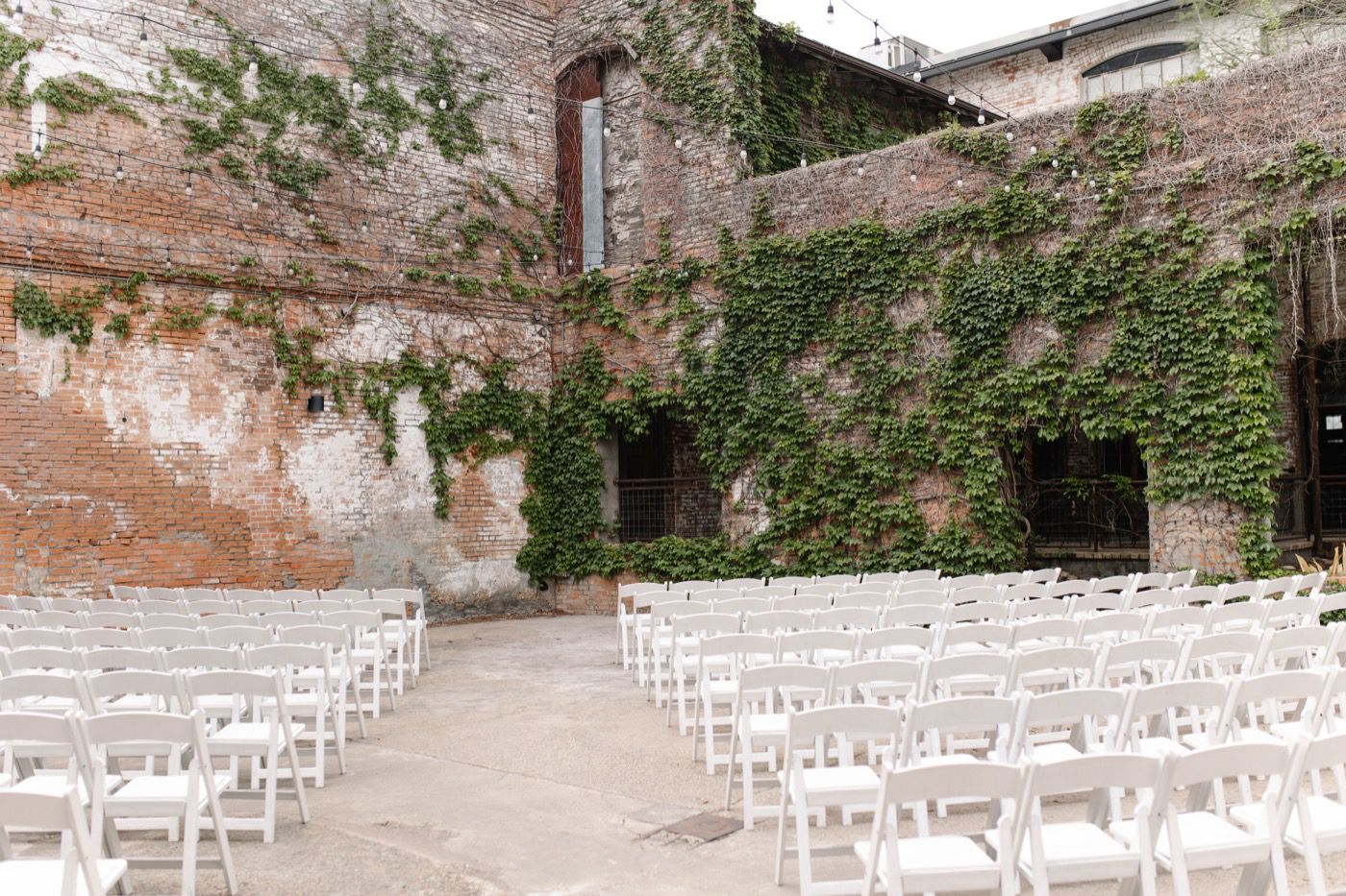 Rows of white folding chairs set up for a ceremony in a brick courtyard covered in ivy and string lights
