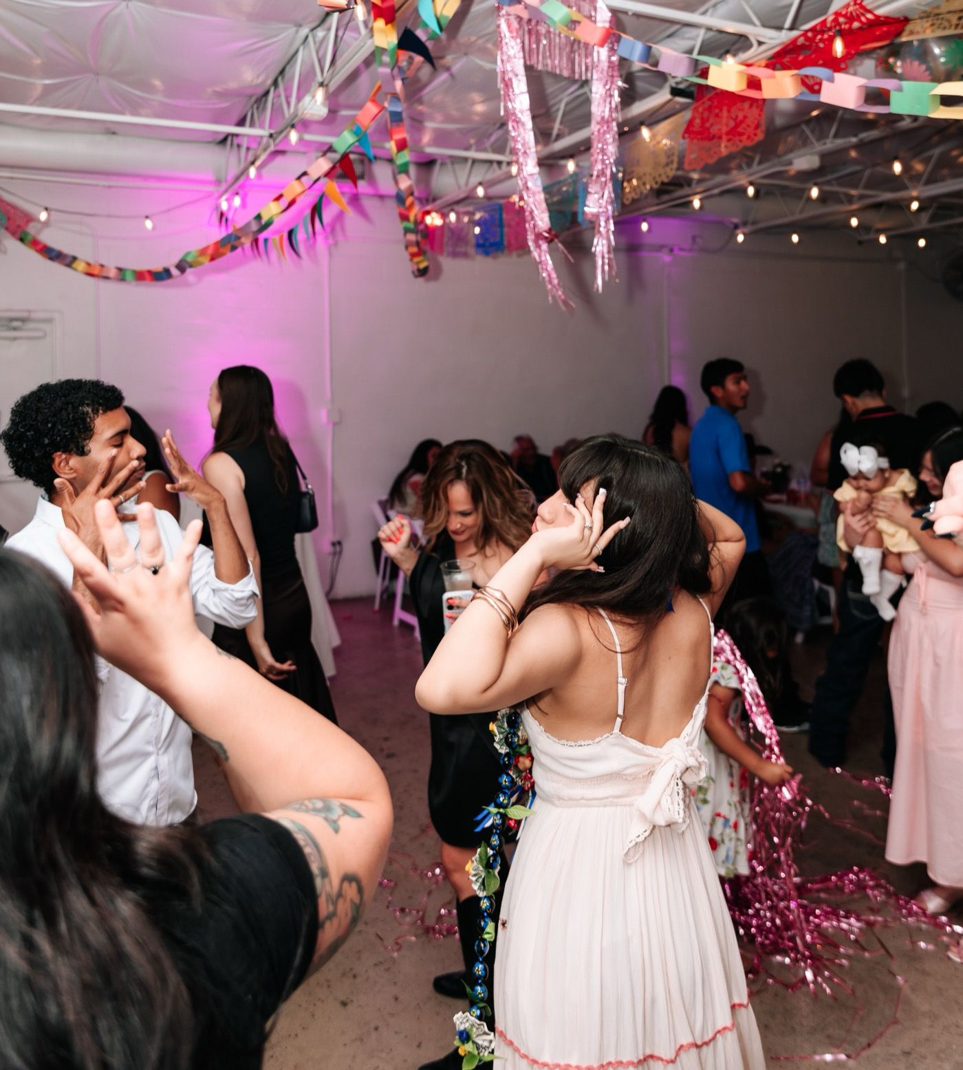 Guests dancing at a birthday party under colorful paper banners and pink tinsel, lit in magenta