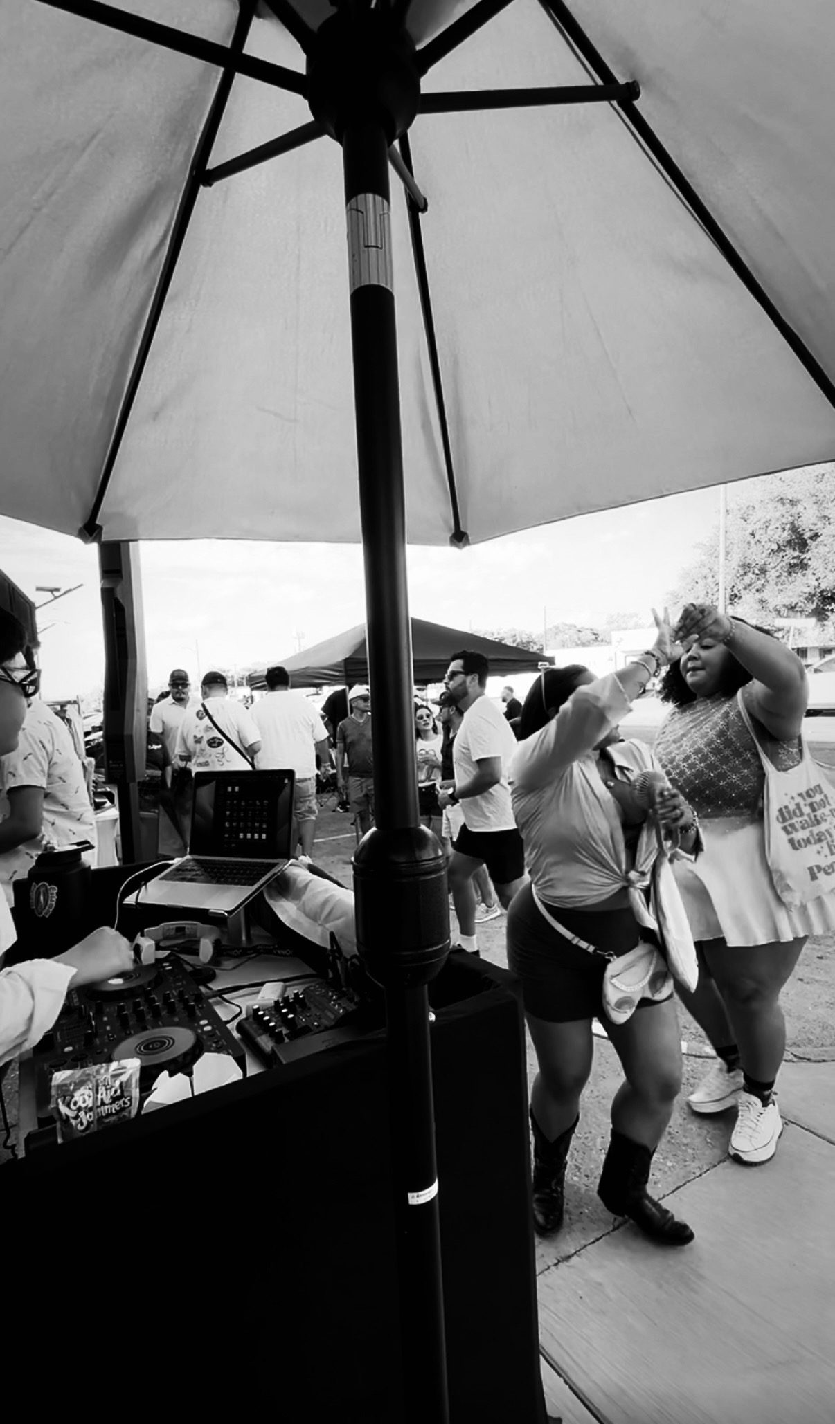 Black-and-white photo of a DJ at the decks under a patio umbrella while two people dance nearby