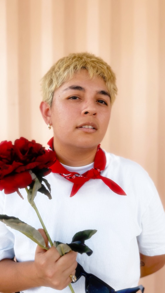 DJ Bri J holding a deep red carnation against a sun-bleached fluted terracotta wall
